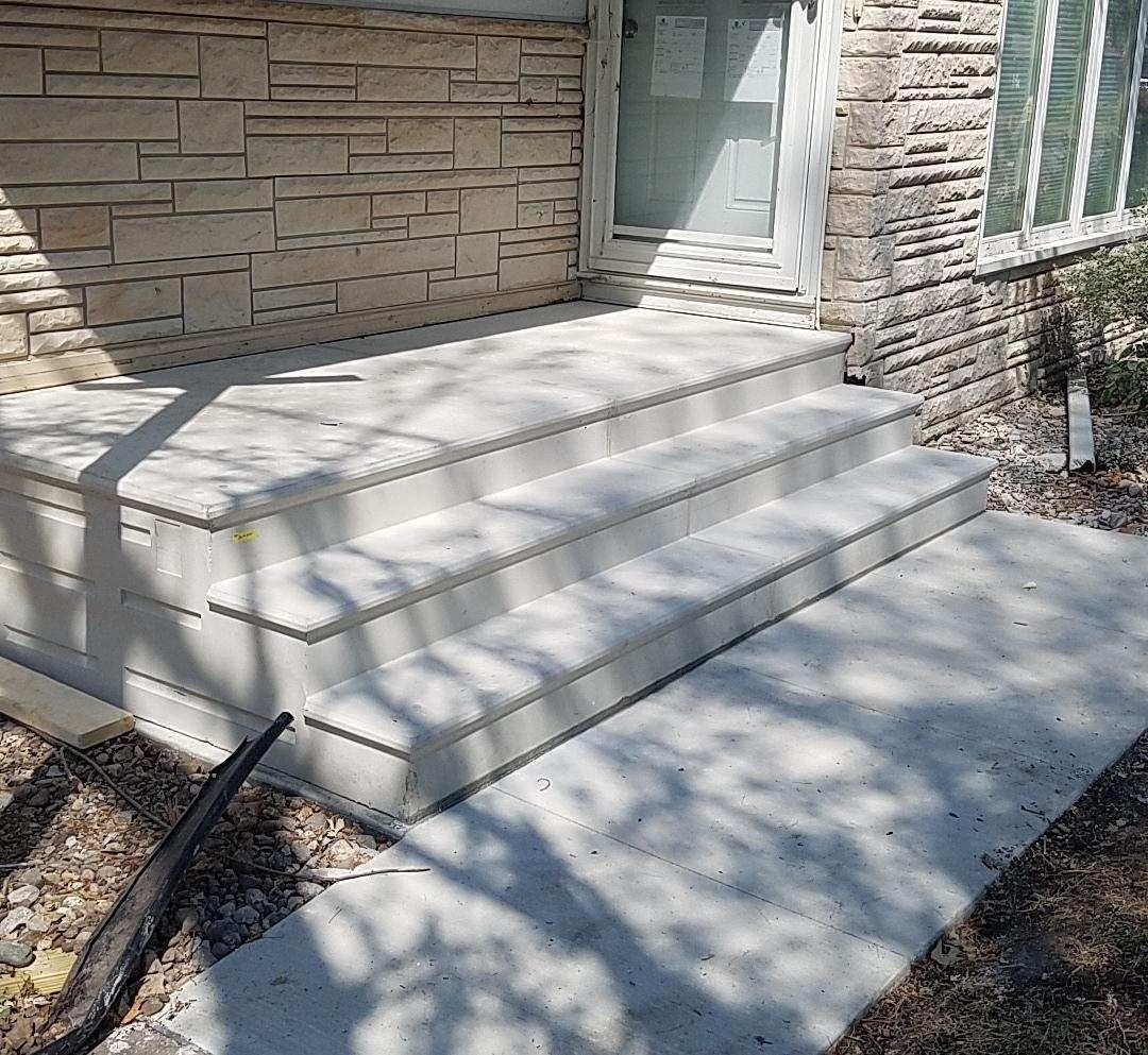 Concrete front steps leading to a stone house entrance, with sunlight and shadows across the walkway.
