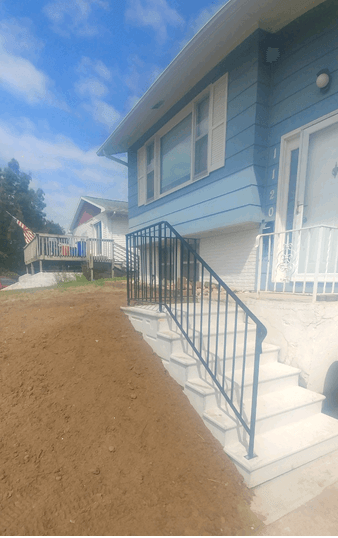 Blue house exterior with white stairs and black railing on a dirt lot under a cloudy sky