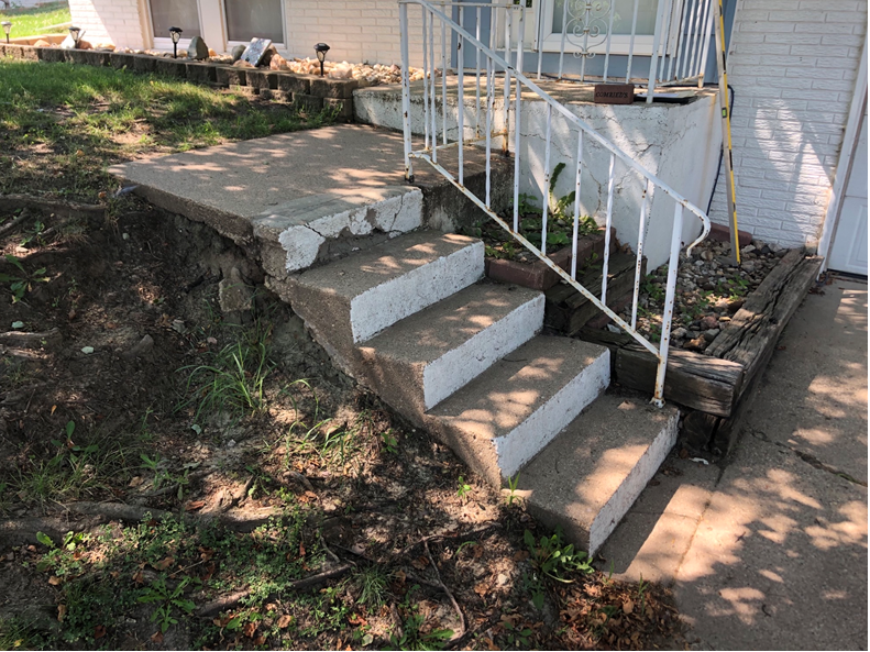 Concrete steps with white railing leading to a house porch, beside a dirt slope and walkway.