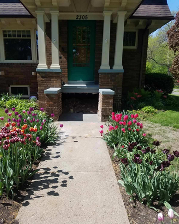 Front porch with green door, brick columns, and a concrete path lined with colorful flowers.