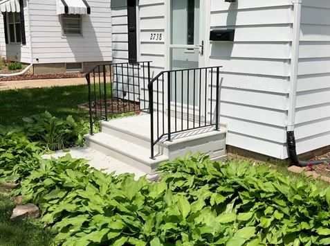 Front porch entrance with gray siding, black railings, and concrete steps beside green plants.
