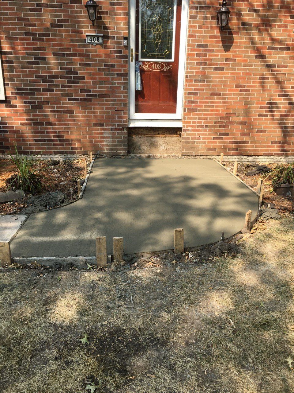 Concrete front stoop leading to a brick house door, with a small retaining edge and dry grass in front.