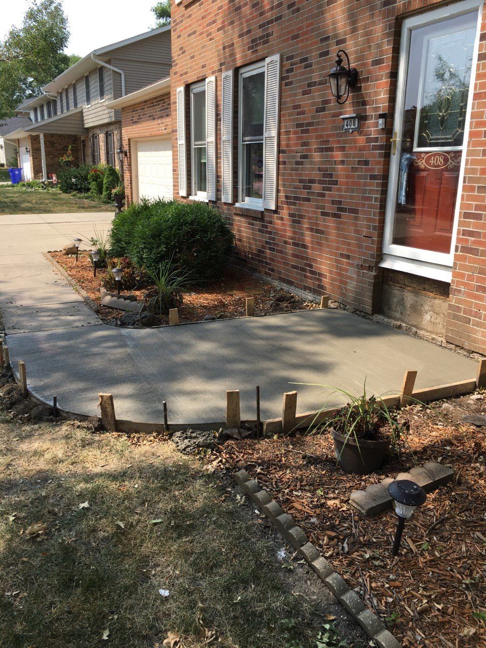 Brick house with a concrete patio, shrub, and small wooden stakes bordering a landscaped bed.