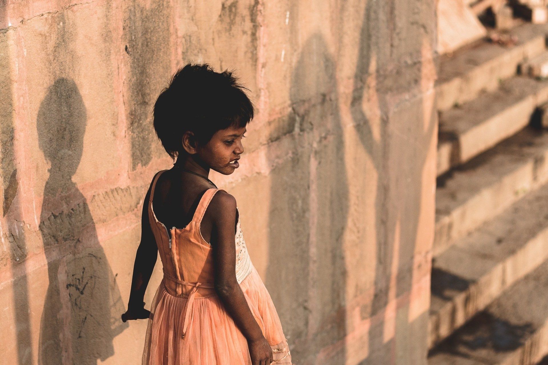 A little girl in a pink dress is standing next to a wall.