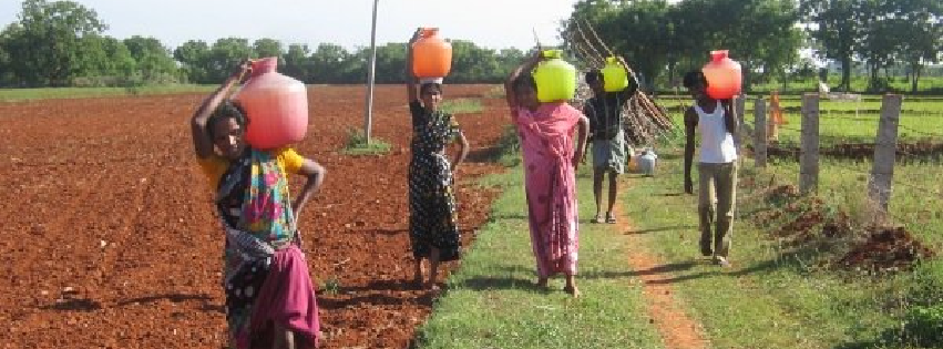 A group of people are walking down a dirt road carrying buckets on their heads.