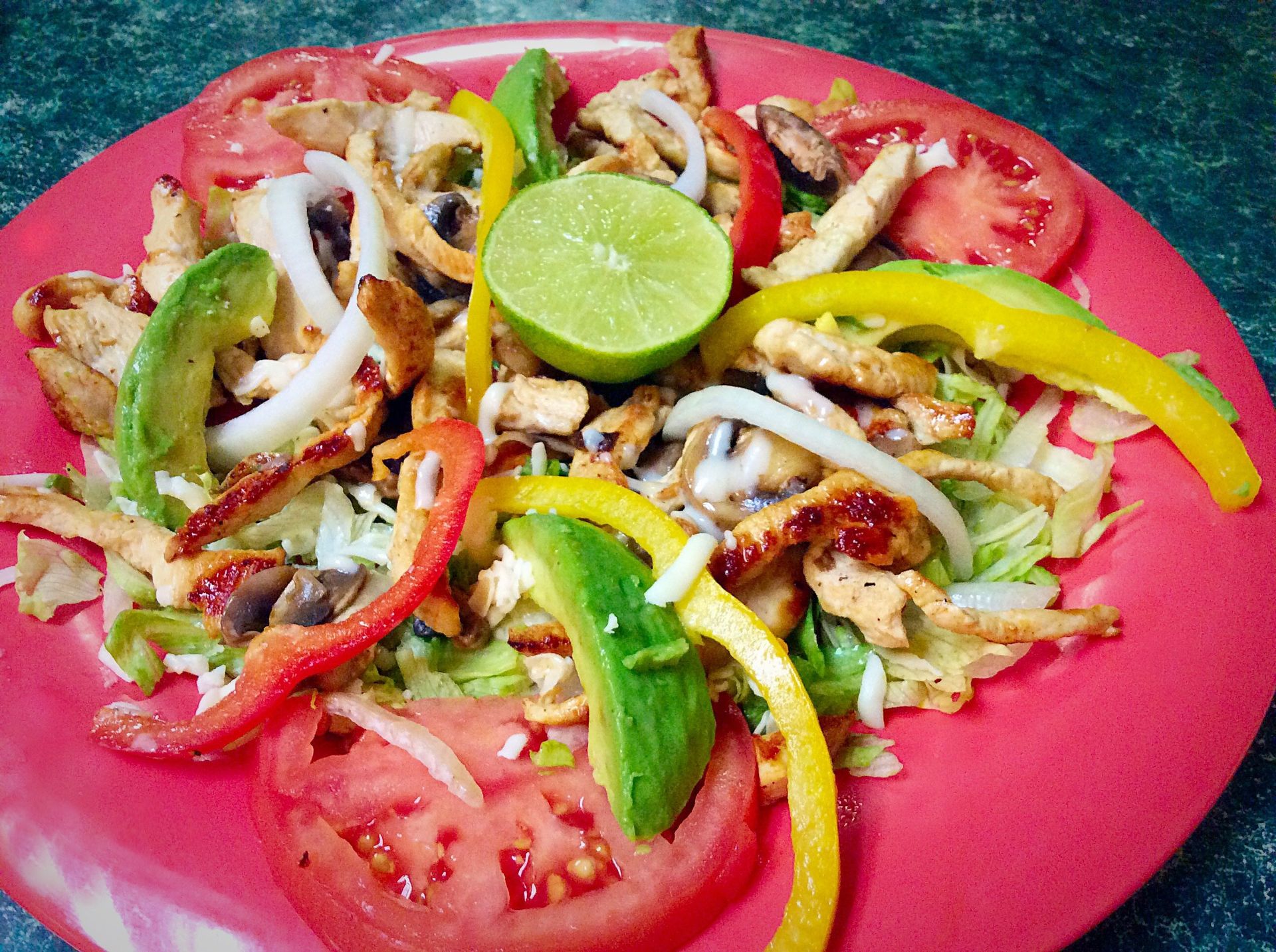 Chicken fajita salad with lime, bell peppers, tomatoes, avocado, and onions on a red plate.