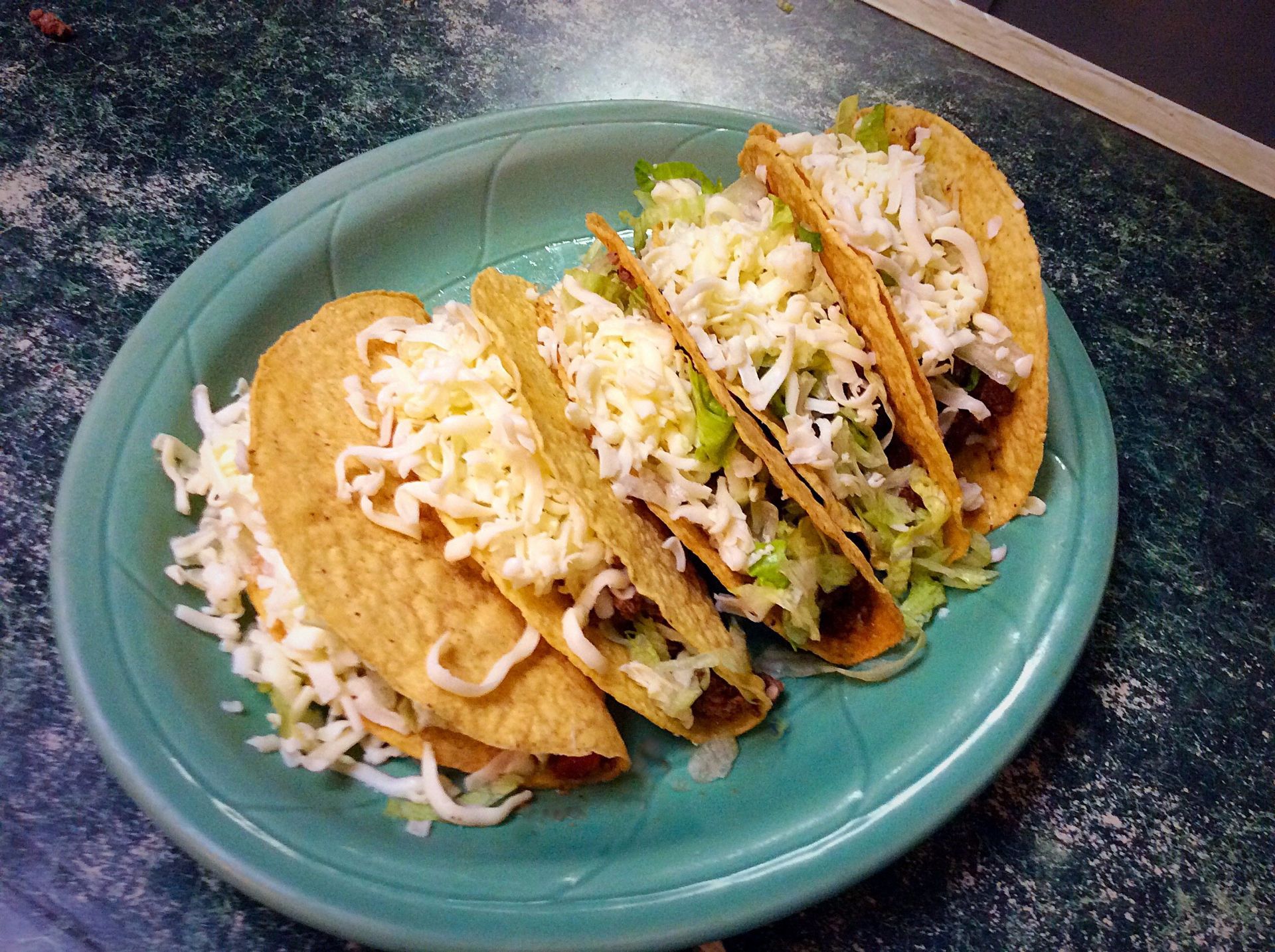 Tacos on a green plate, filled with lettuce and topped with shredded cheese.