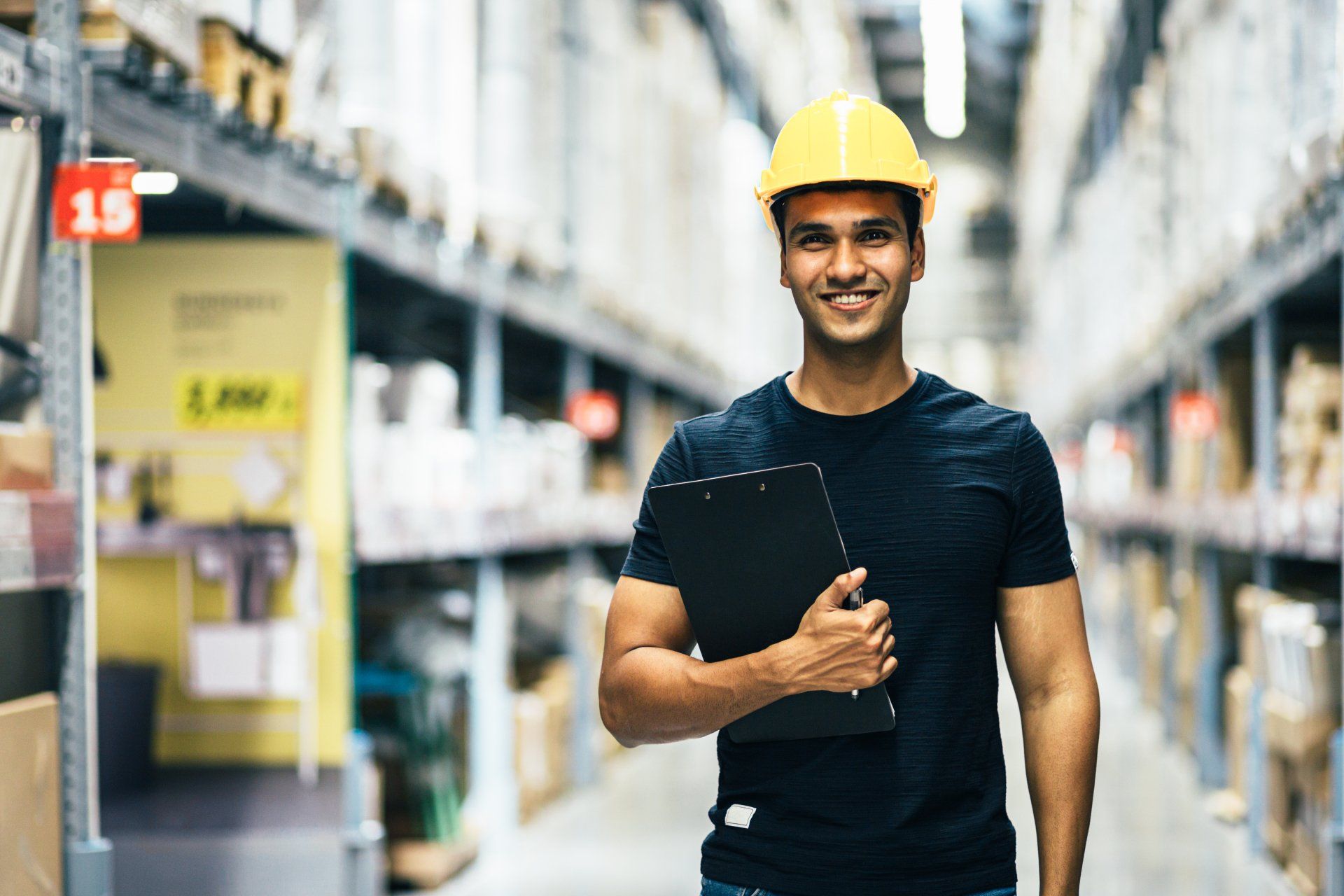 A man wearing a hard hat is holding a clipboard in a warehouse.