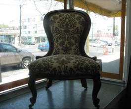 Upholstered antique chair with dark wood frame and patterned fabric, in a window with a city street view.