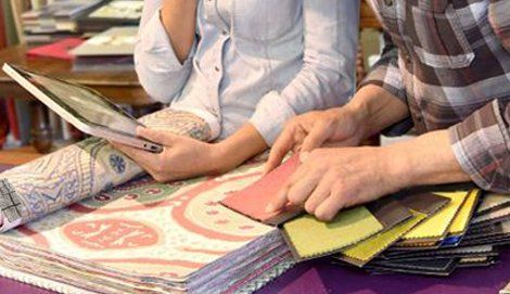Person holding a tablet and another person examining fabric swatches. A rug sample is in the foreground.