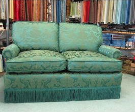 Teal damask loveseat with fringed skirt, set against shelves of fabric bolts.