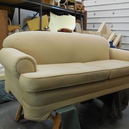 Cream-colored loveseat in a workshop; it's on sawhorses. The couch has rolled arms, a skirt, and two cushions.