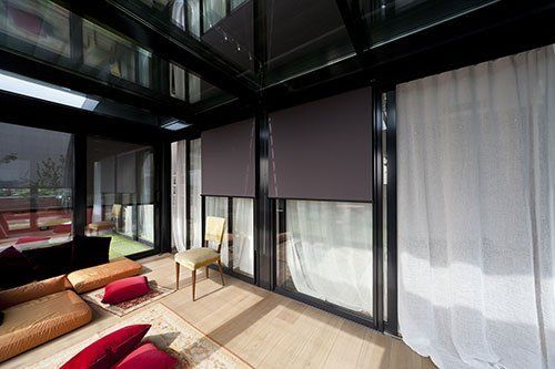 una veranda con una sedia, dei cuscini su un pavimento in legno e delle vetrate con le tende di color bianco
