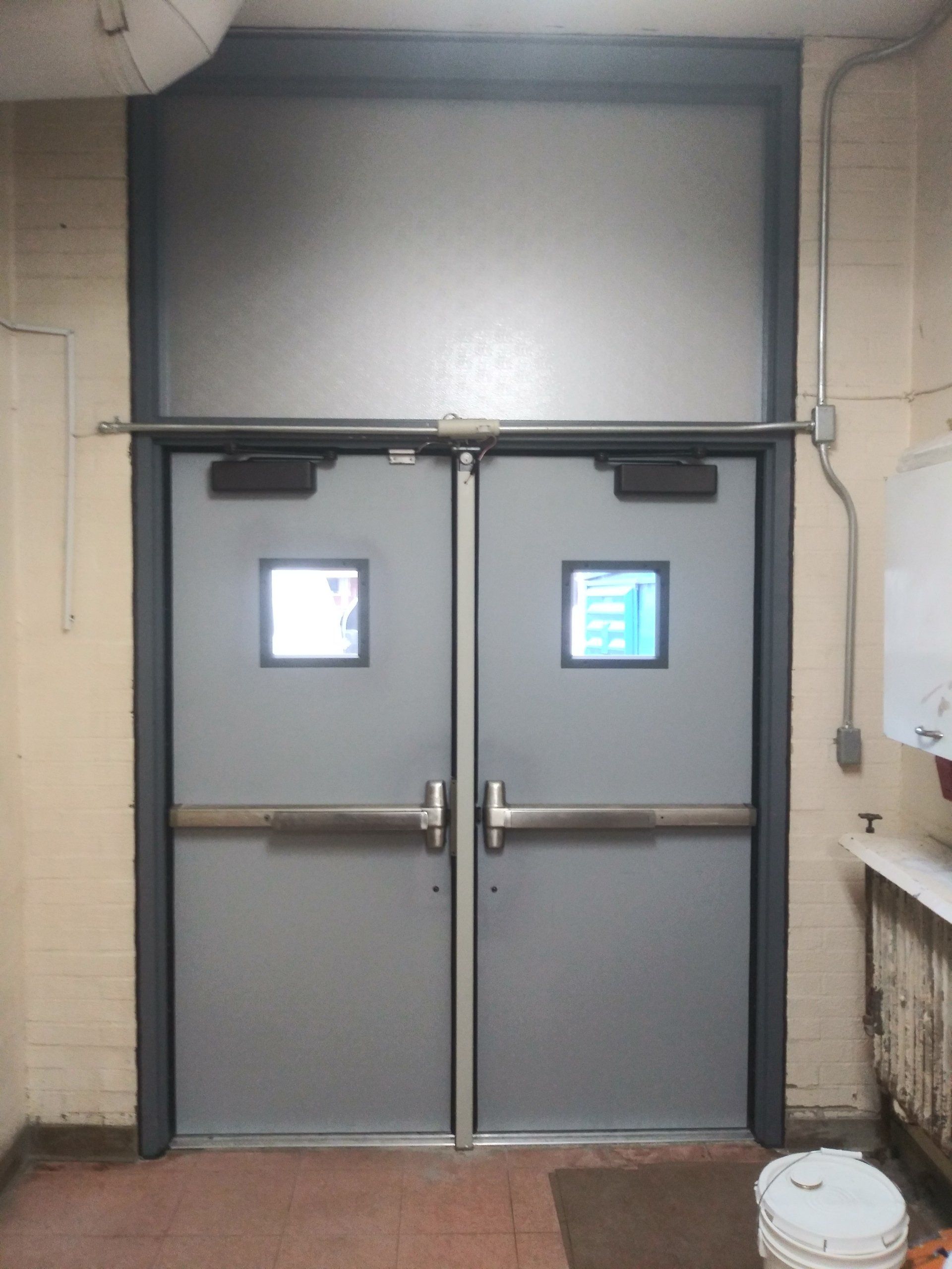 Gray double industrial doors with small windows in a narrow hallway, each with a push bar.