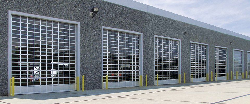 Exterior view of a building with multiple transparent garage doors. Gray building with yellow bollards.