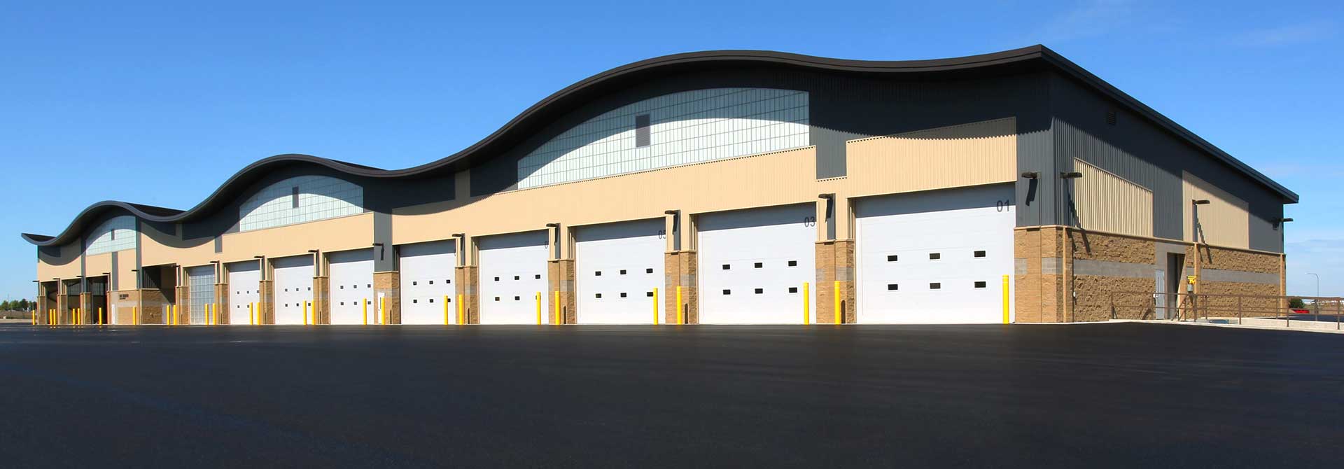 Wide industrial building with a curved roof and several white garage doors on a paved lot.
