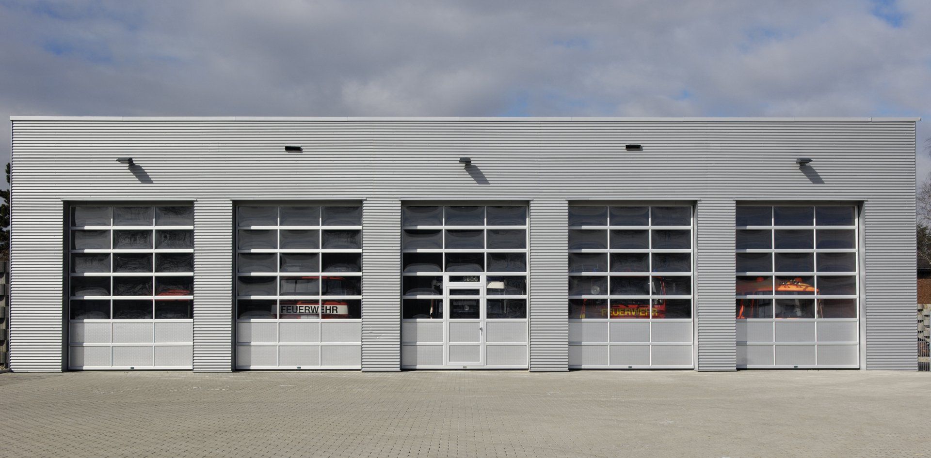 A modern gray building with four garage doors, each with glass panes. Cloudy sky above.
