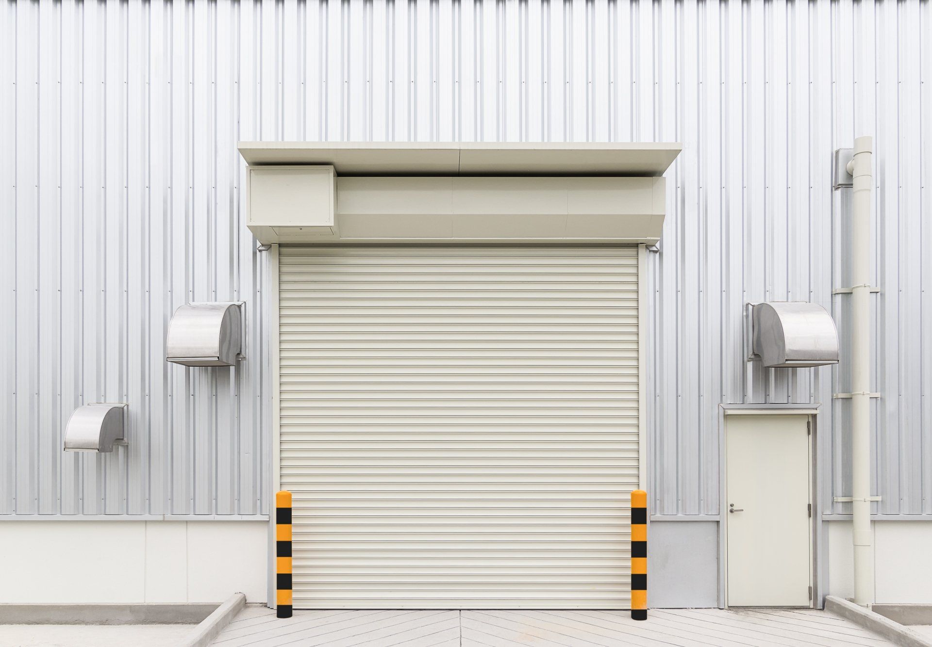 A closed, beige roll-up door on a silver industrial building with a small white door and air vents.
