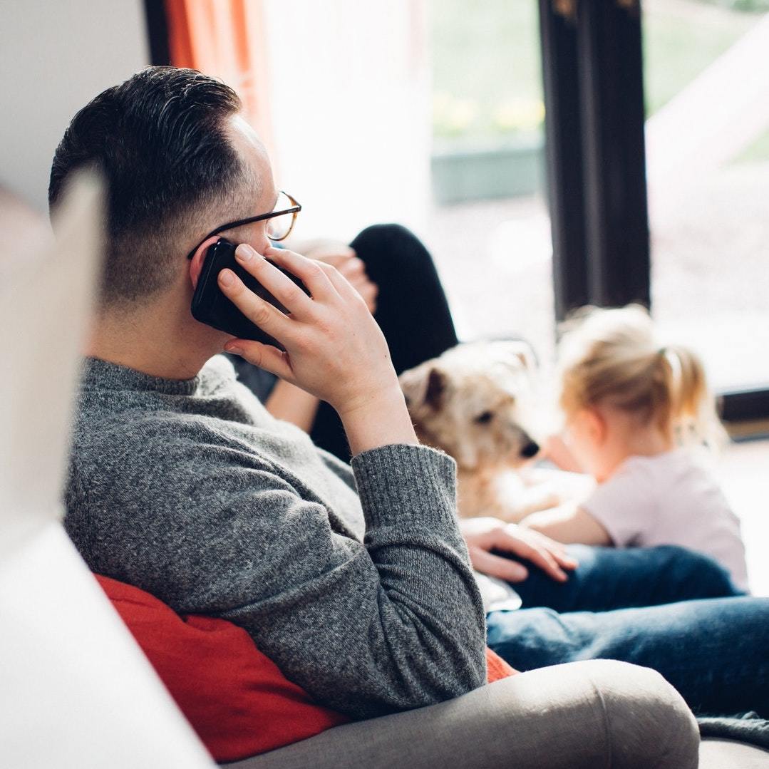 father talking on phone with dog and daughter in background