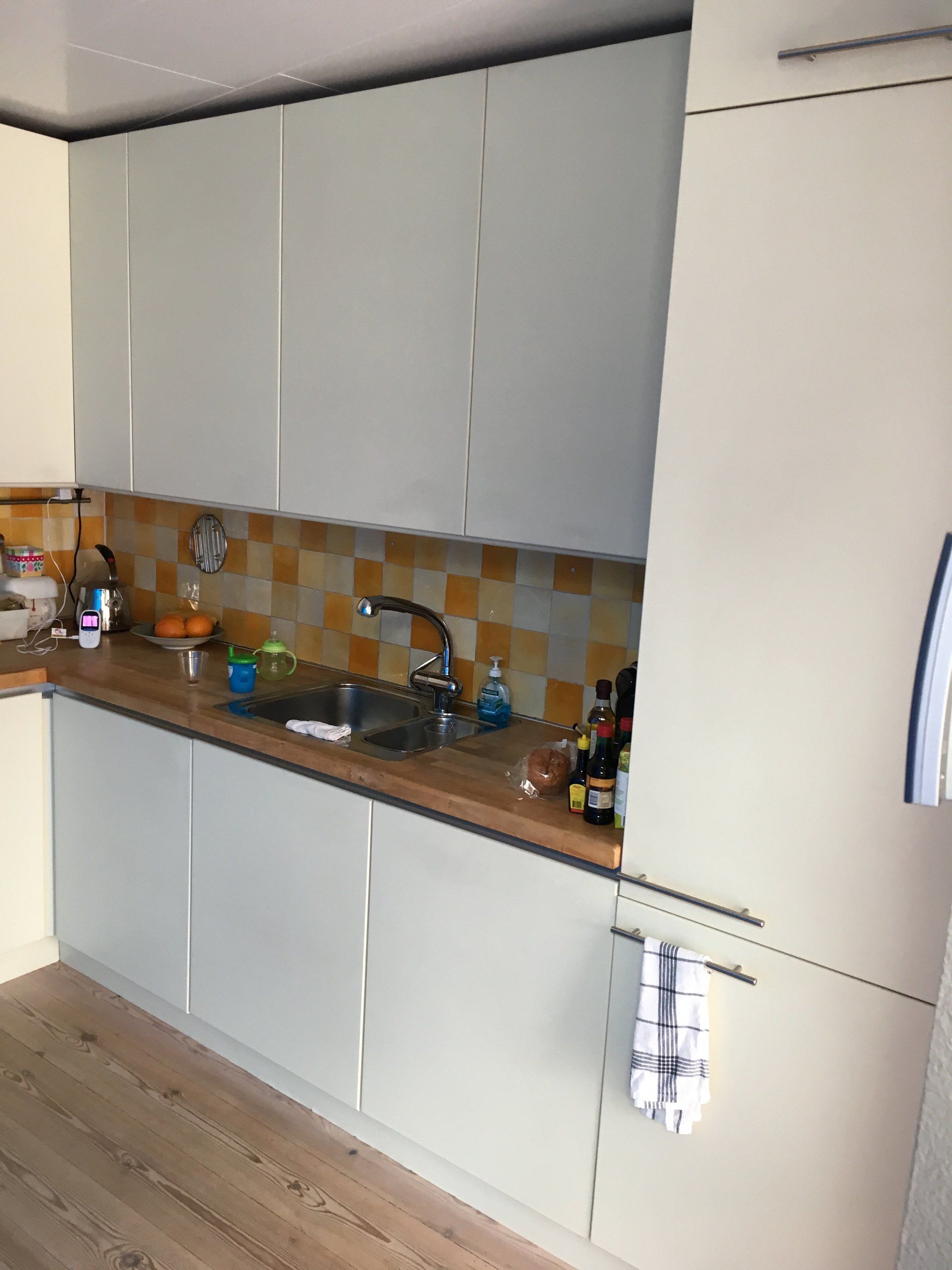 Moderne keuken met witte kasten, bruin aanrechtblad, betegelde achterwand, spoelbak en koelkast.