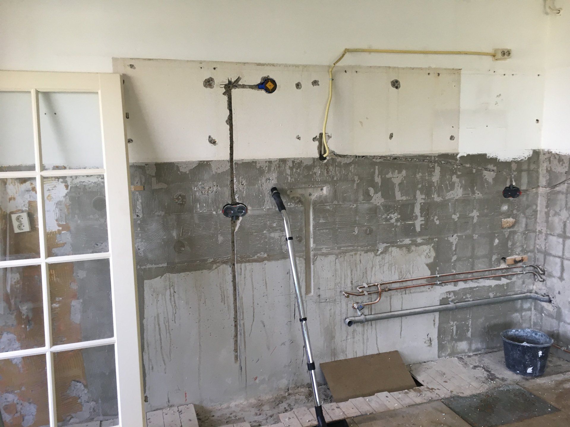 Keukenrenovatie met onafgewerkte grijze stucwanden, zichtbare leidingen en een deur aan de linkerkant.