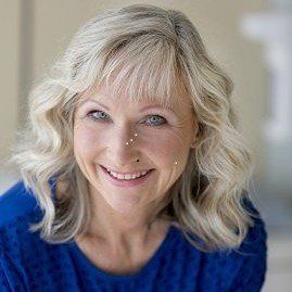 Eine Frau mit blonden Haaren trägt einen blauen Pullover und lächelt.
