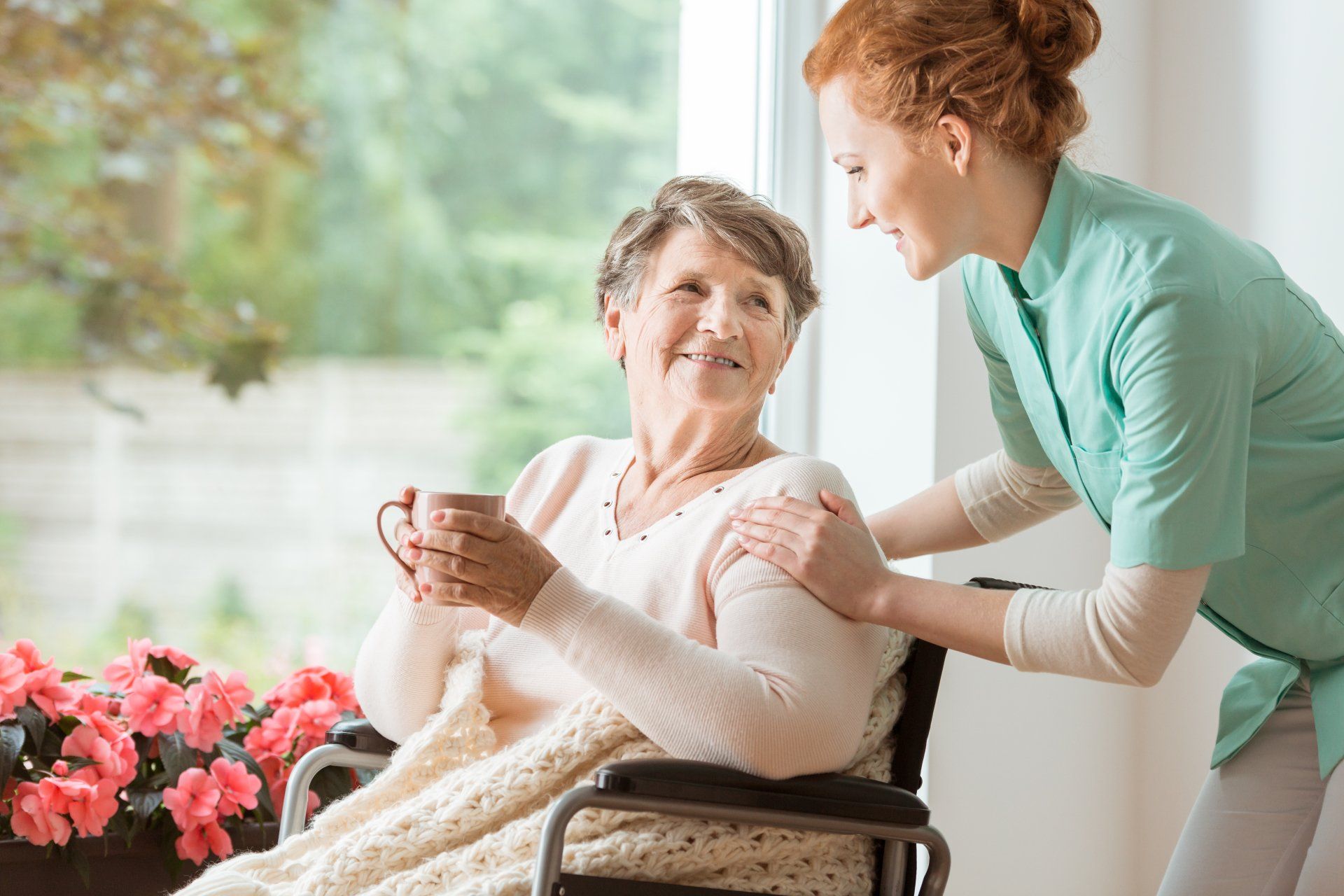 An elderly woman in a wheelchair is holding a cup of coffee and talking to a nurse.