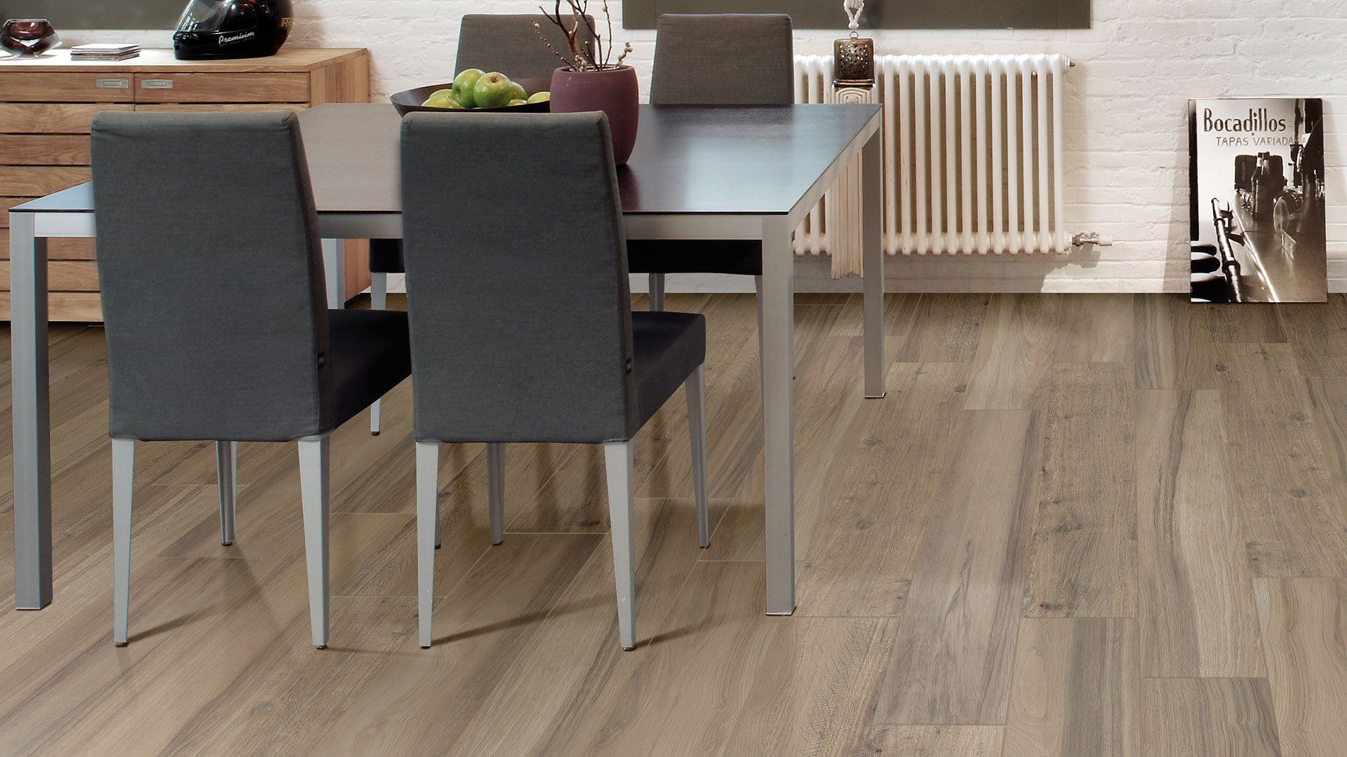 Sala da pranzo moderna con tavolo in legno, due sedie scure e pavimento in legno chiaro vicino a un termosifone.