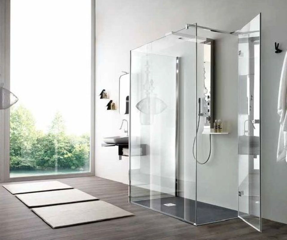 Bagno moderno e minimalista con box doccia in vetro, lavabo a parete e ampia finestra con vista sul verde.