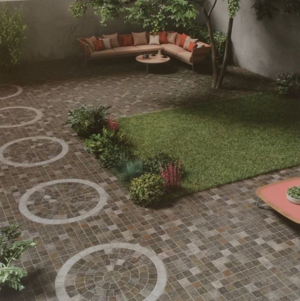 Piccolo patio paesaggistico con pavimentazione in mattoni, motivi circolari in pietra, prato, arbusti e una panchina esterna curva.