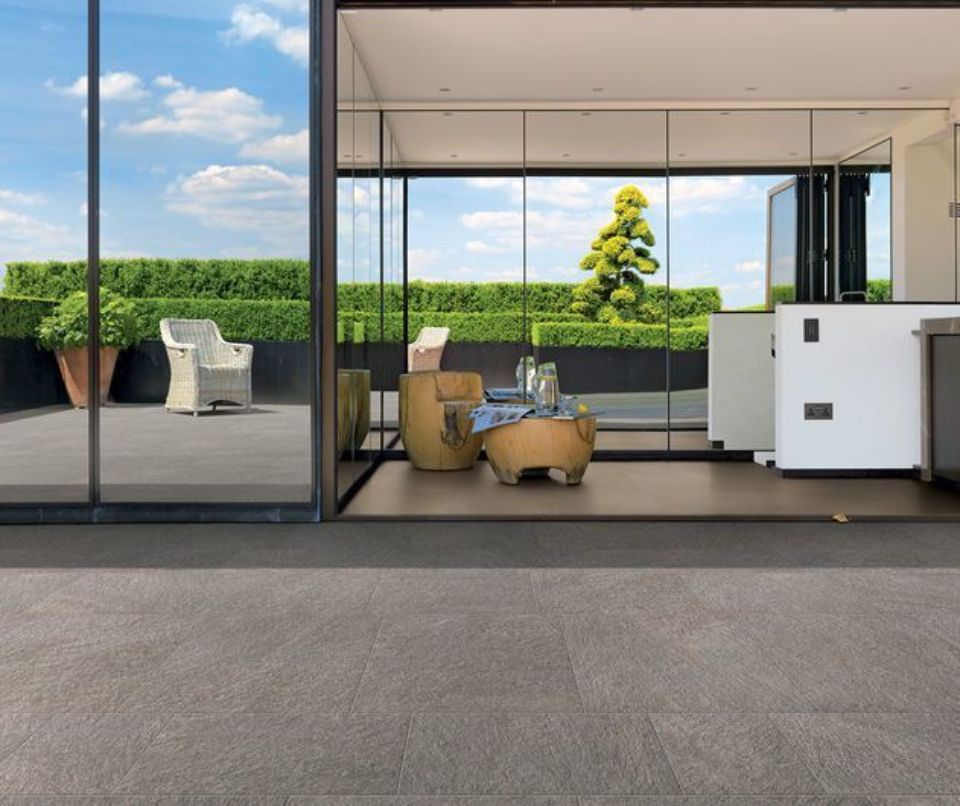 Patio moderno con pareti in vetro, sedie in vimini e piante in vaso, affacciato su un giardino di siepi ben curato.