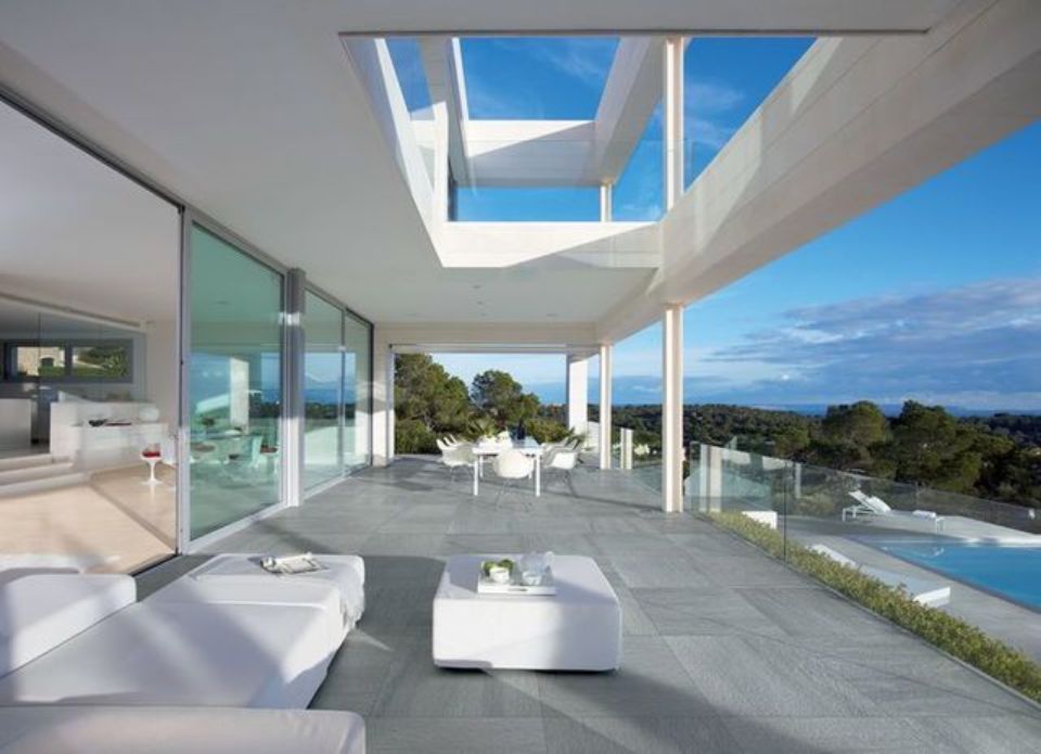 Terrazza di una villa moderna bianca con vista sull'oceano, pareti in vetro, lettini prendisole e cielo azzurro.