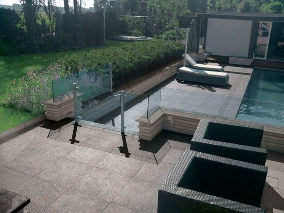 Patio moderno con piscina, lettini da esterno e ringhiera in vetro adiacente al giardino.