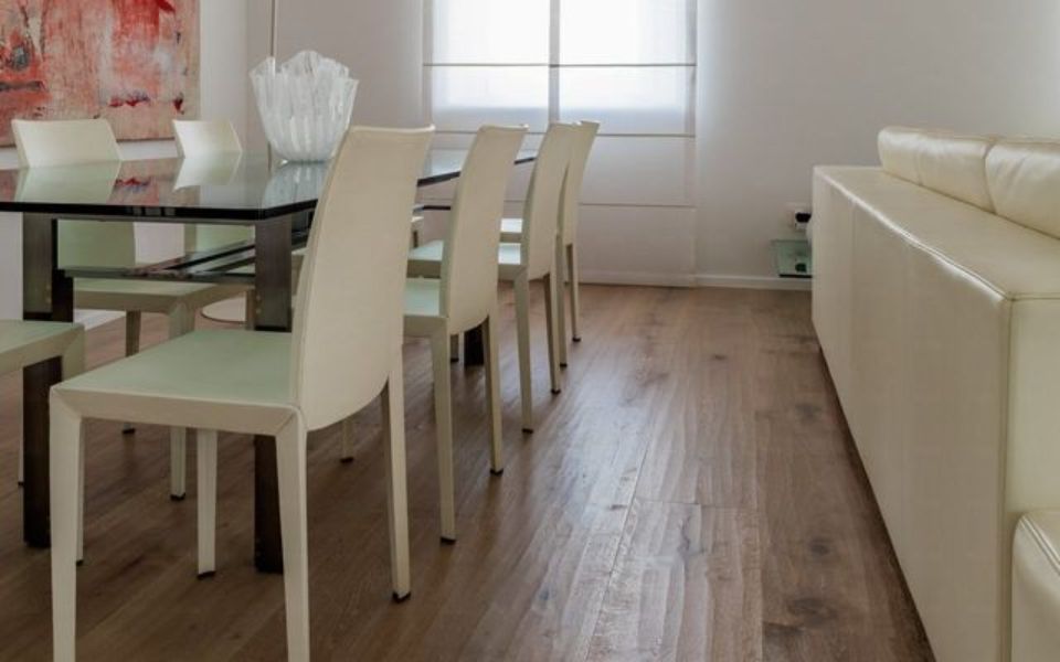 Sala da pranzo moderna con sedie bianche, tavolo in vetro, divano color crema e pavimento in legno.