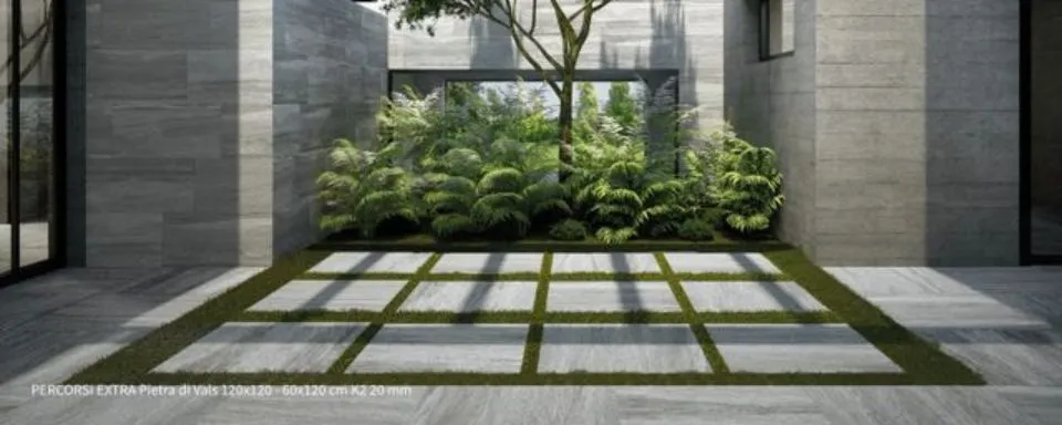Cortile moderno con muri in pietra, piante verdi rigogliose e un'ombra a griglia illuminata dal sole sul pavimento piastrellato.