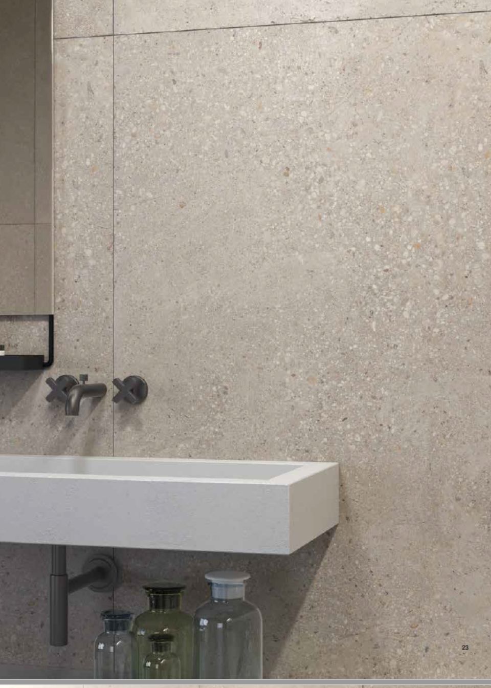 Lavabo del bagno con rubinetti a parete, piastrelle beige e flaconi riposti al di sotto.