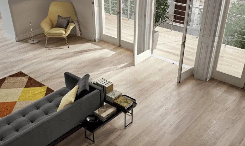 Luminoso soggiorno con poltrona beige, divano grigio, tappeto colorato e porte finestre che si aprono su un balcone.