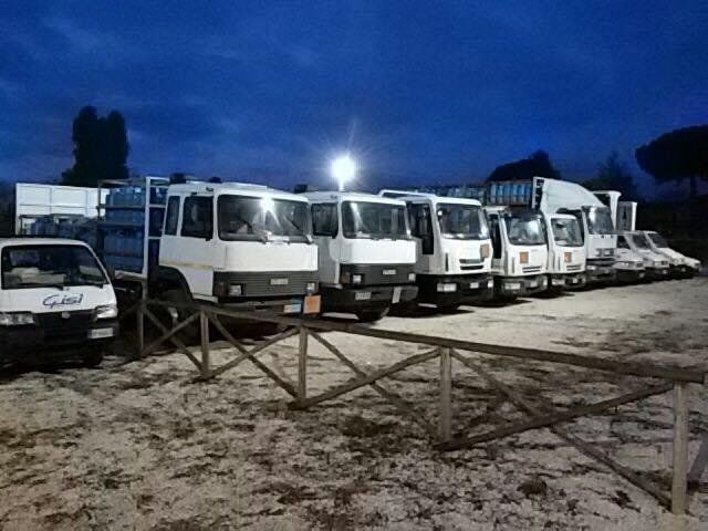Una fila di furgoni bianchi parcheggiati in un piazzale sabbioso al crepuscolo, illuminati da un faro luminoso.
