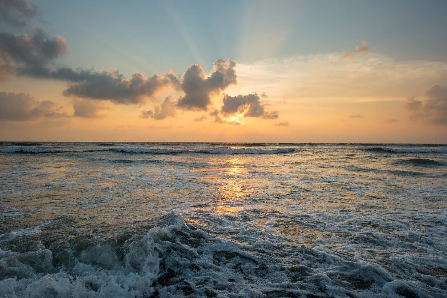 Sunset over ocean; waves crashing. Orange and gold sky, reflecting on water.