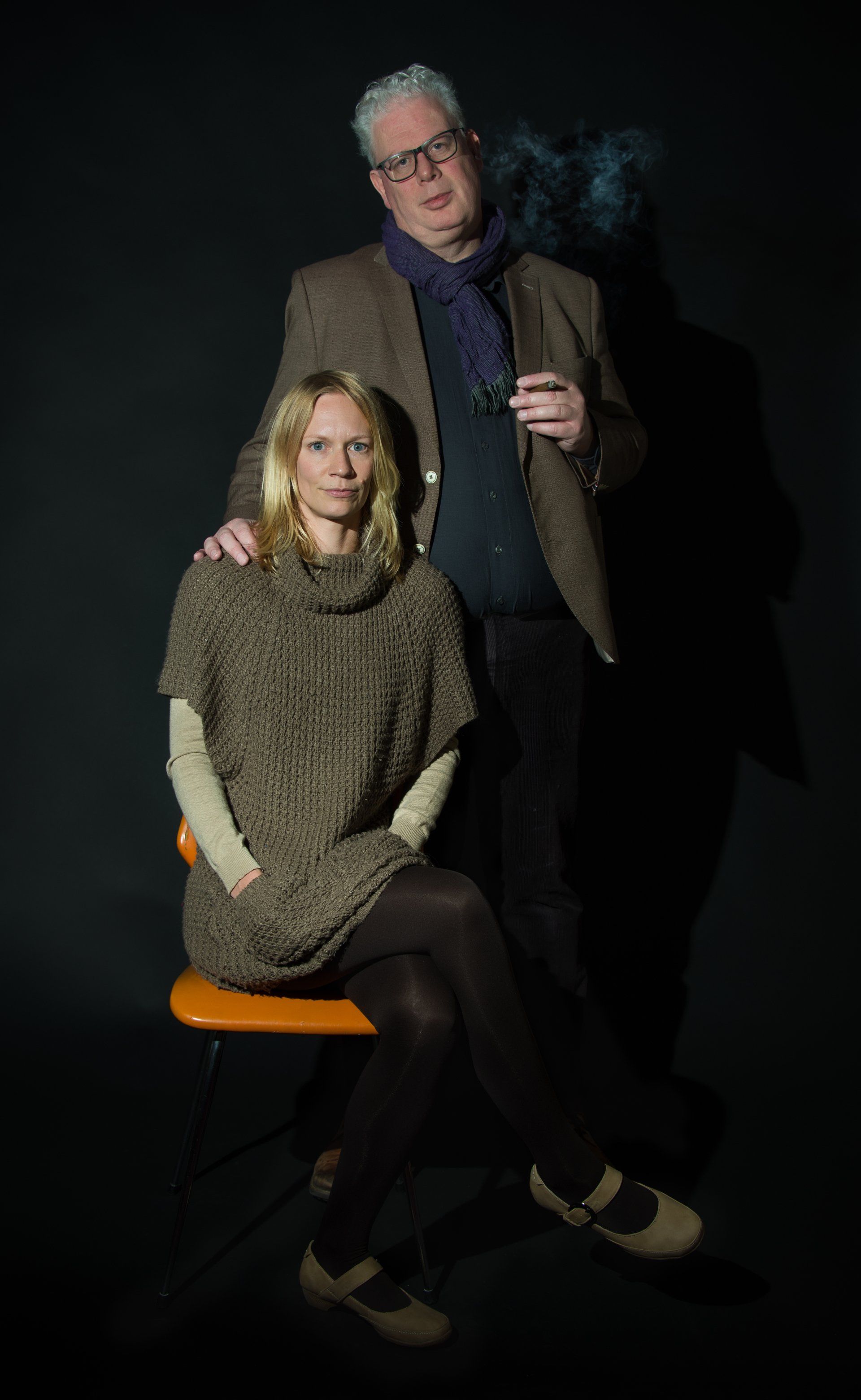 Two people posing in a dark studio, one seated on an orange stool and one standing behind, both facing the camera.