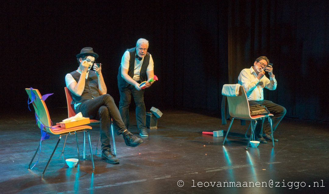 Three performers on chairs in a dark stage scene, one standing between two seated musicians.