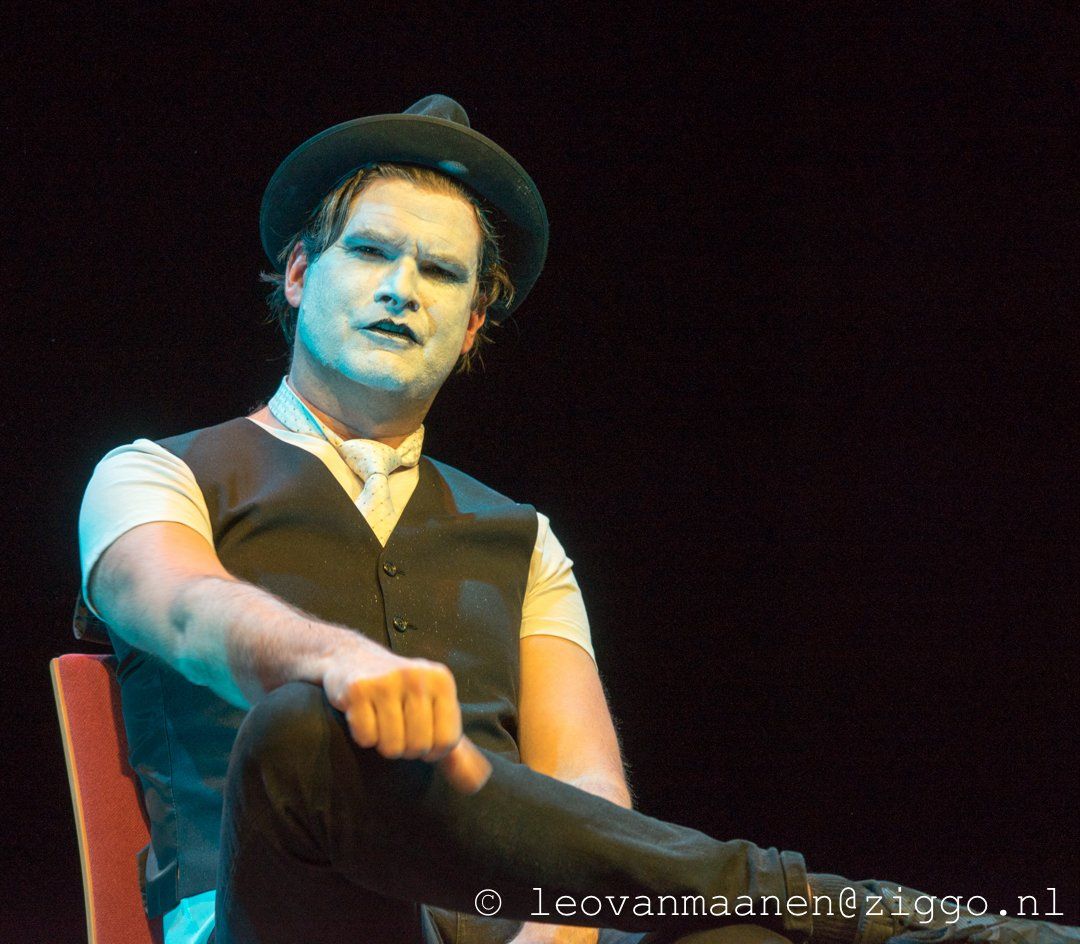 Man in a hat and vest onstage, pointing forward under blue lighting.