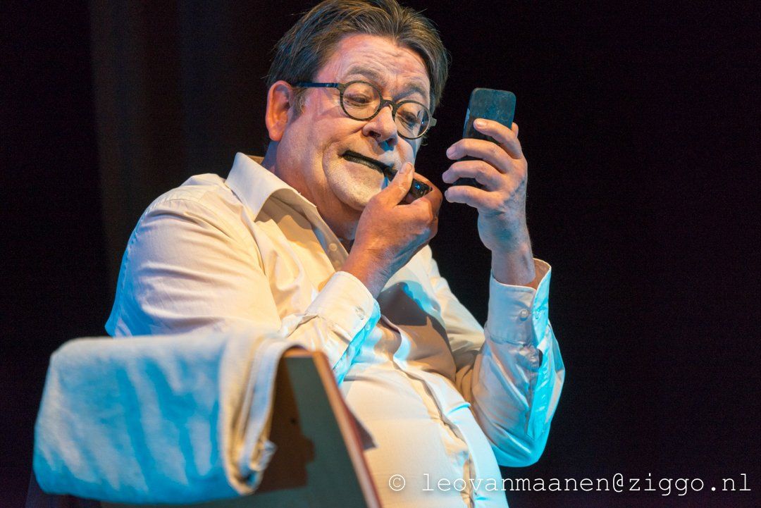 Man on stage holding a smartphone, speaking into a microphone beside a white draped chair