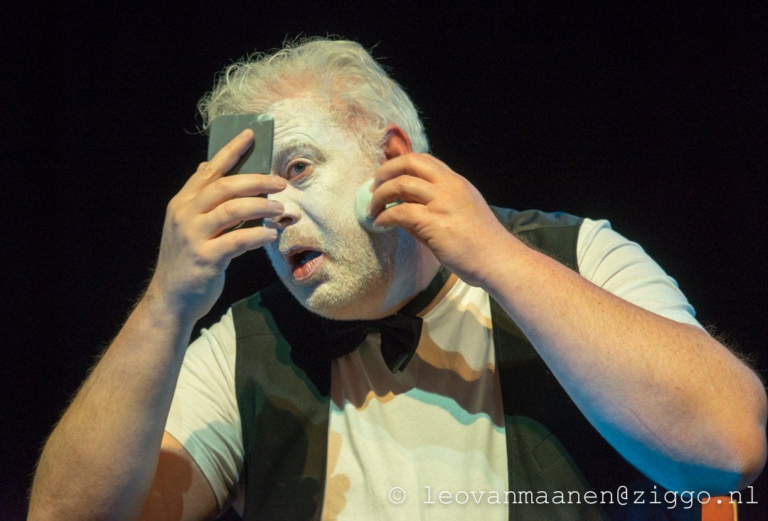 Man with white face paint holds his head in his hands on a dark stage.