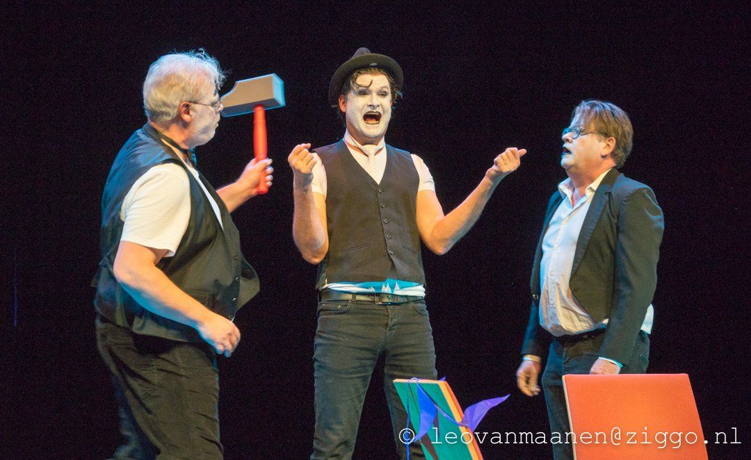 Three actors on a dark stage, with one holding a hammer and the center actor gesturing animatedly.
