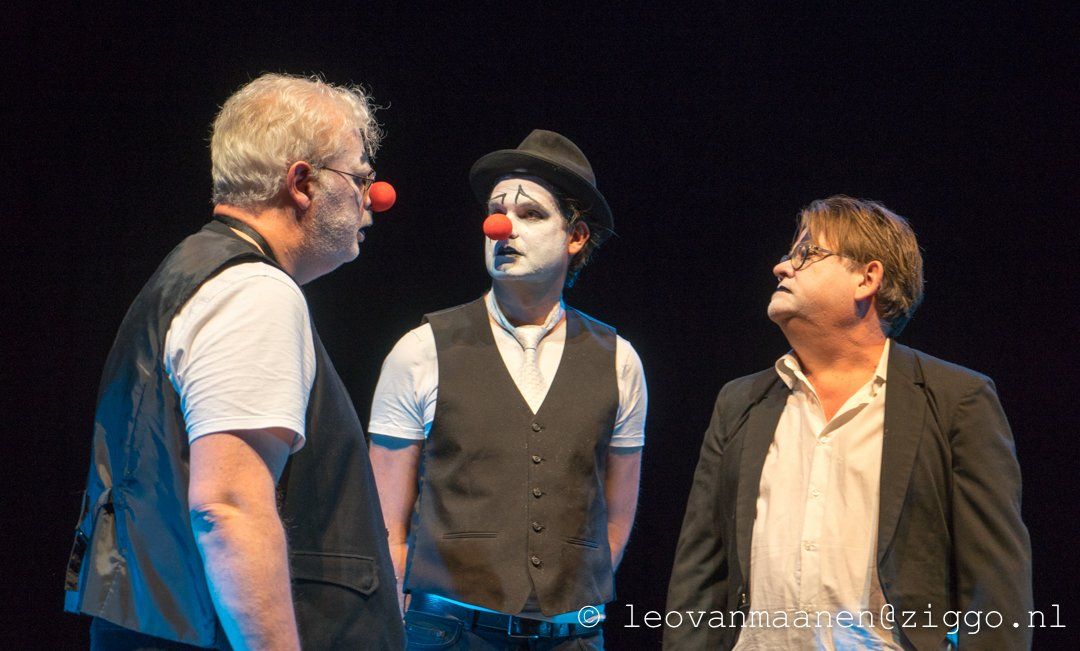 Three performers onstage, including a clown with red nose, facing each other in a dark theater scene