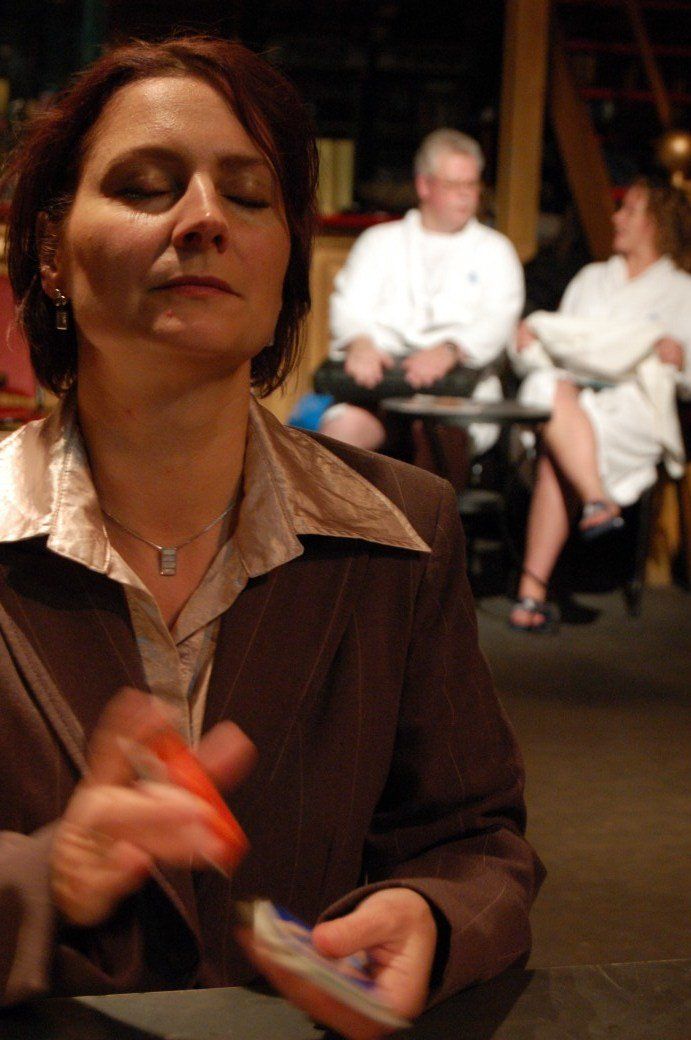 Woman in brown jacket gesturing in foreground, with two people seated softly blurred in the background.