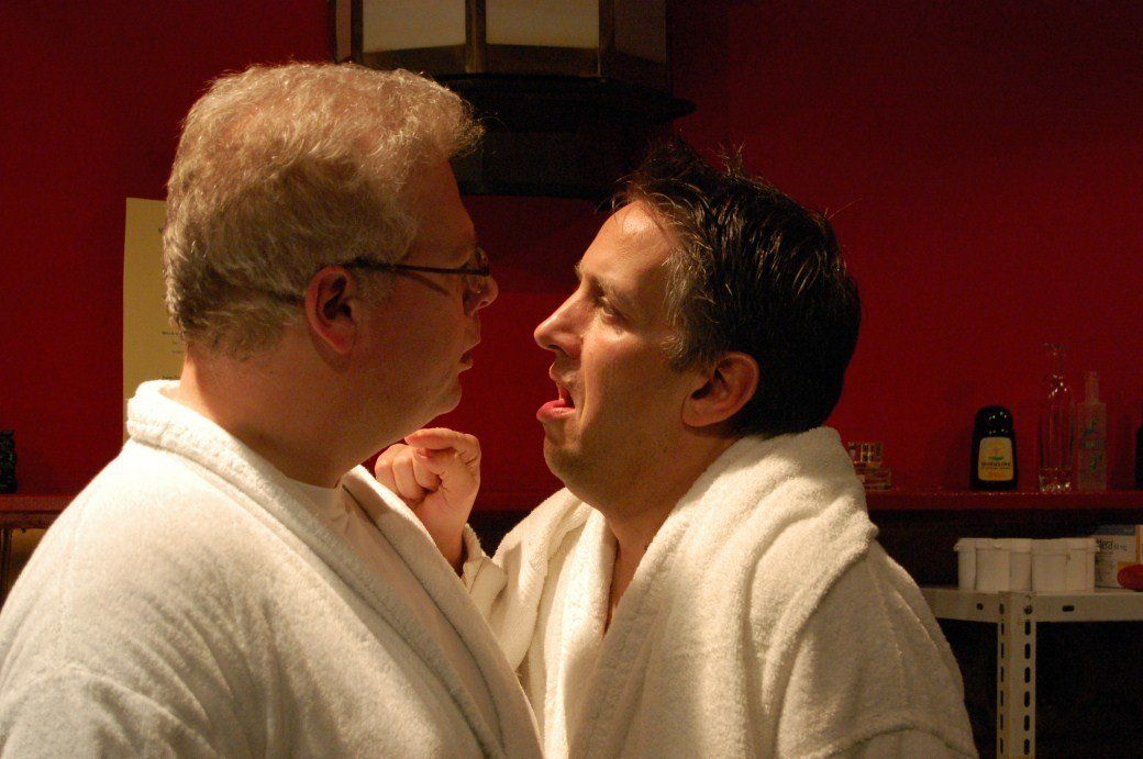 Two men in white robes face each other closely in a dim red room.
