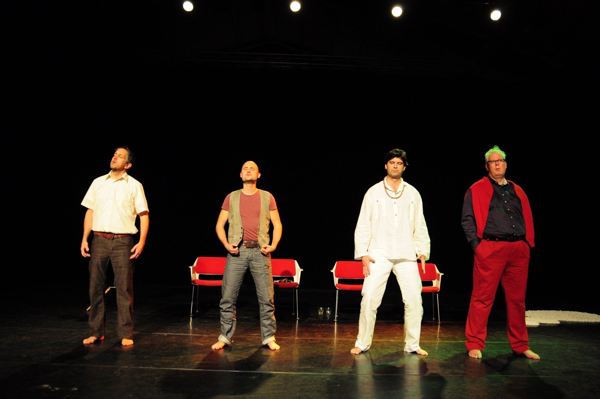 Four performers stand on a dark stage under spotlights, with red chairs behind them.