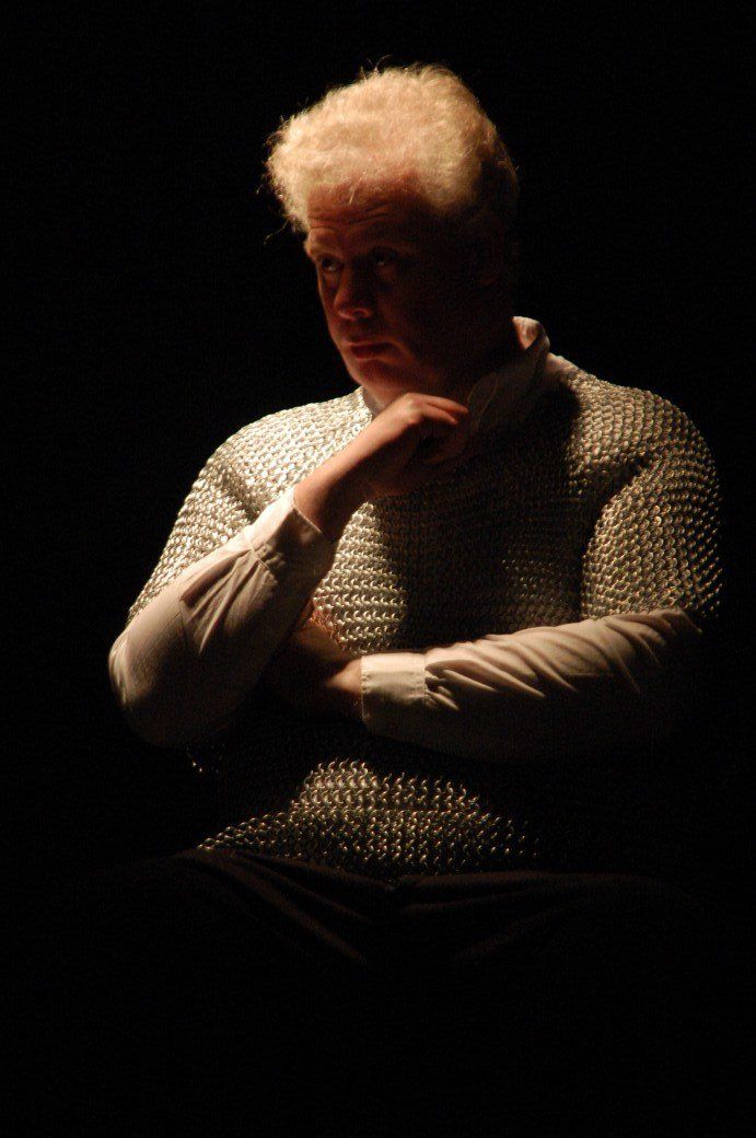 Person seated in dim light, hand at chin, wearing a textured sweater against a dark background